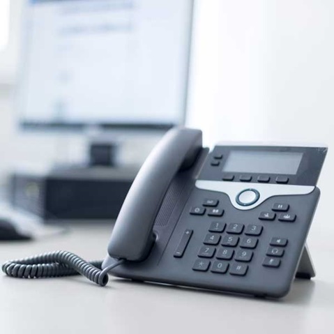 A modern grey office telephone with a keypad, placed on a desk in front of a computer monitor.